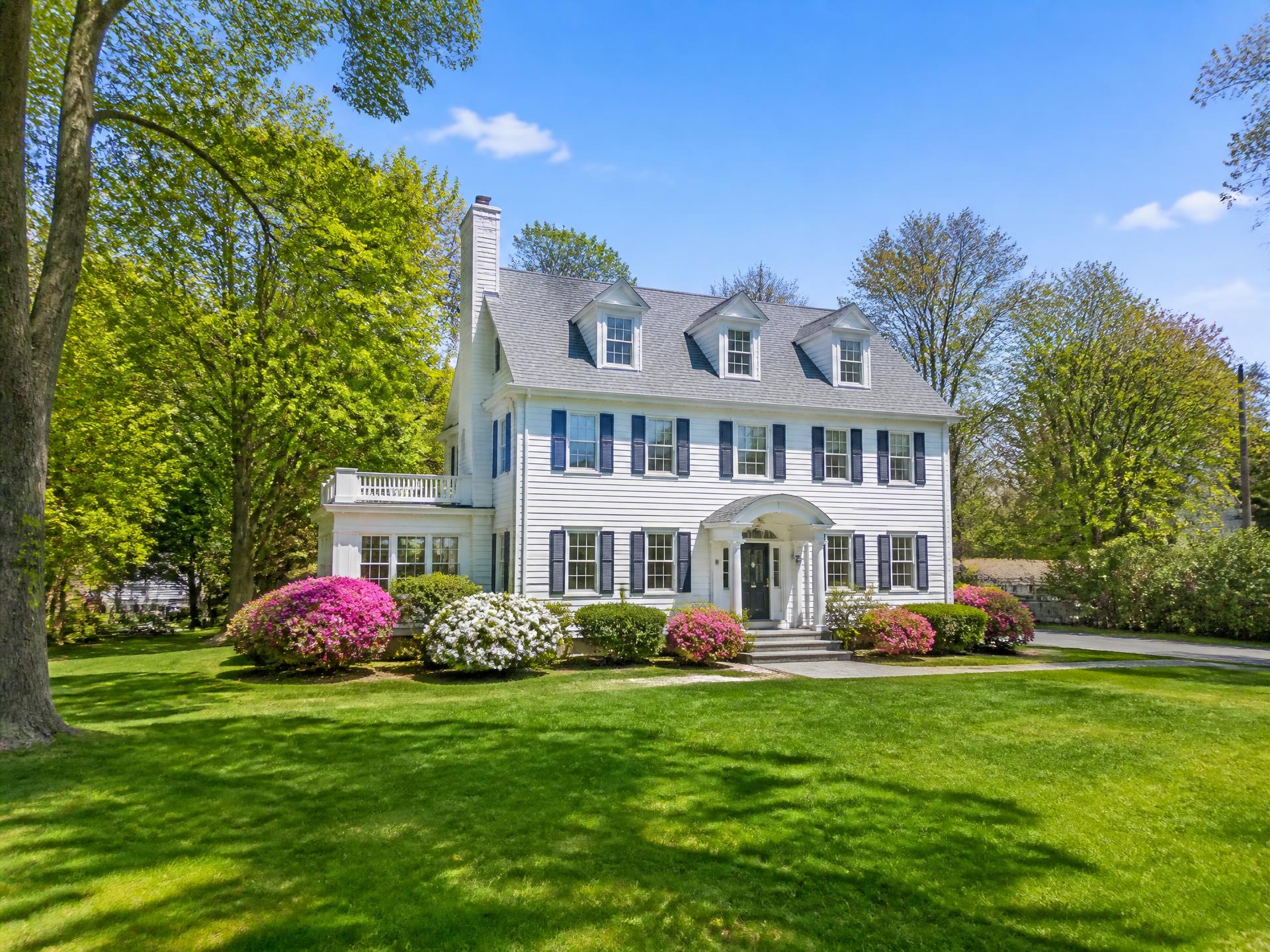 Spacious white New England colonial home with navy shutters, flowering shrubs and mature trees in a sunny Belmont setting